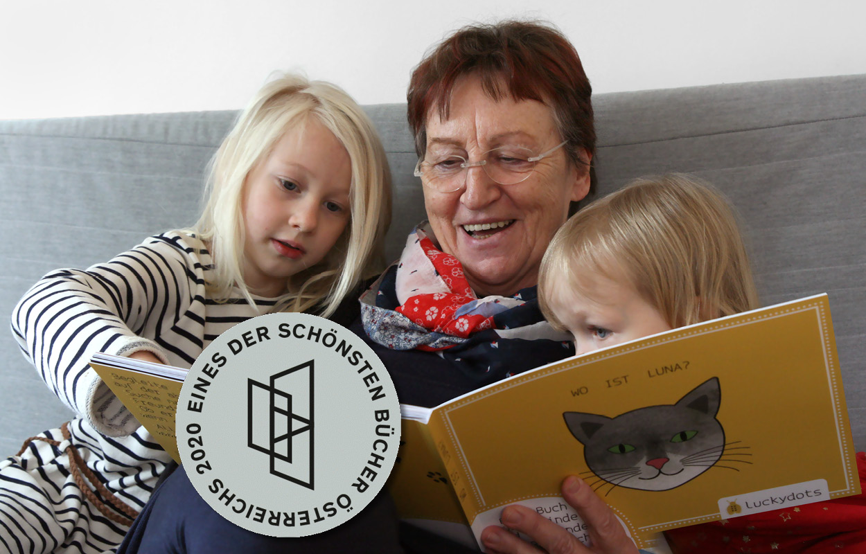 Oma und zwei Mädchen lächeln, als sie das Buch aufschlagen. Logo Schönste Bücher